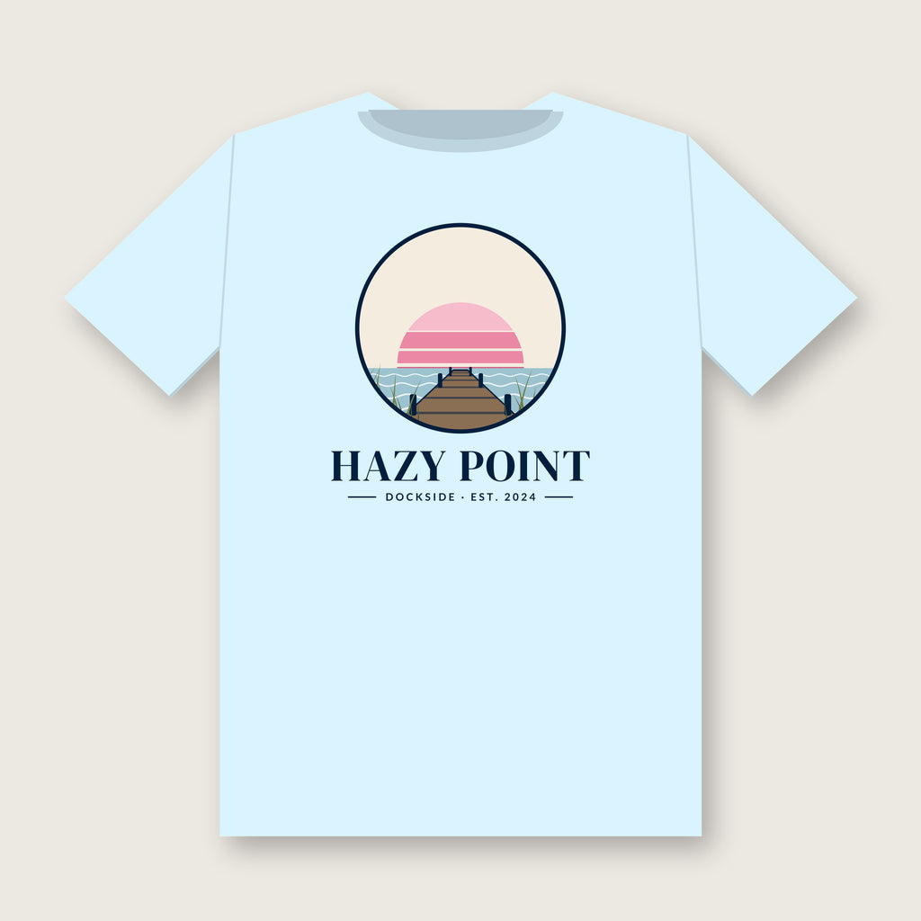Back of the chambray-blue Hazy Point Dock Sunset Tee: a roundel of pink sunset bands, still water and weathered dock planks, with HAZY POINT and Dockside, EST. 2024 beneath